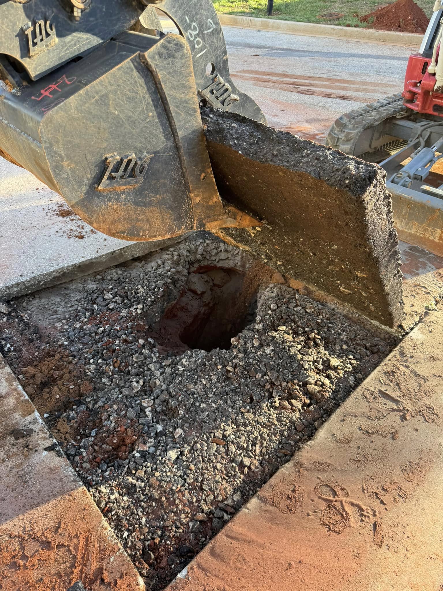 An excavator lifts a square piece of asphalt, revealing a hole in the ground beneath.