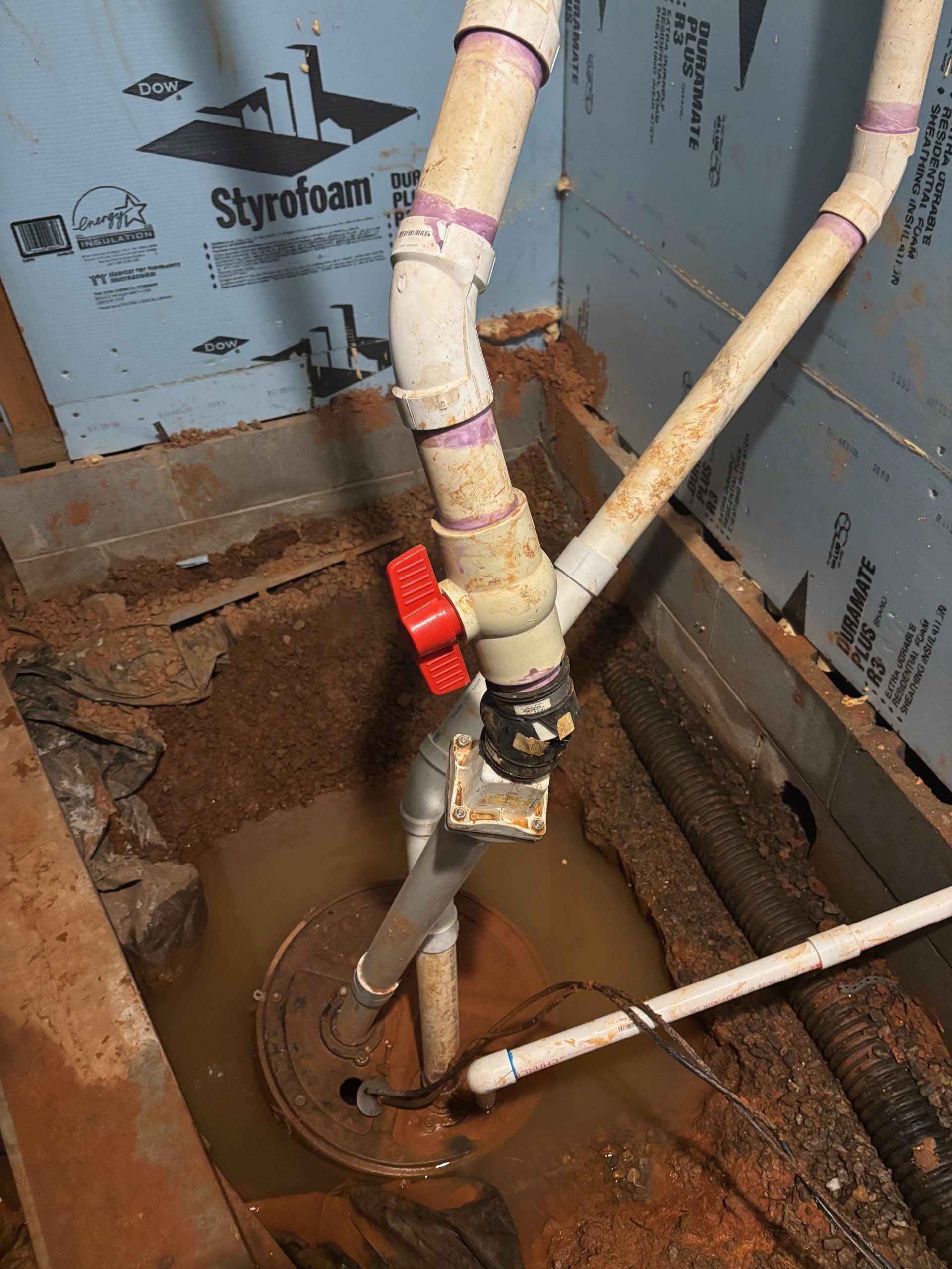 Sump pump installed in a muddy, water-filled basement corner with PVC pipes and a red valve.