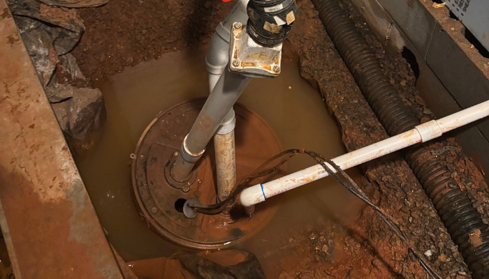 Untitled (700 x 400 px) A sump pump surrounded by muddy water in a flooded basement with exposed pipes and wet ground.