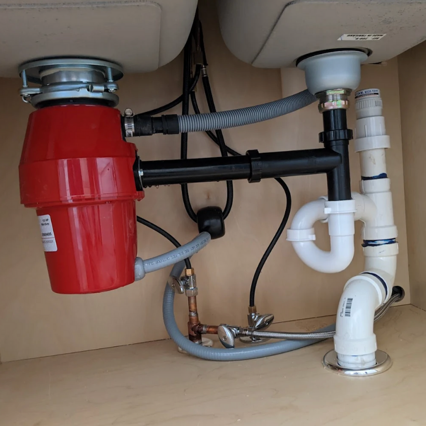 Plumbing under a kitchen sink, showing a red garbage disposal and various pipes and drain connections.