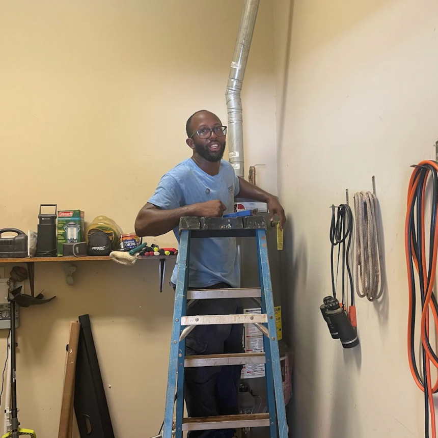 Man standing on a ladder fixing pipes in a workshop with tools and equipment on shelves and walls.