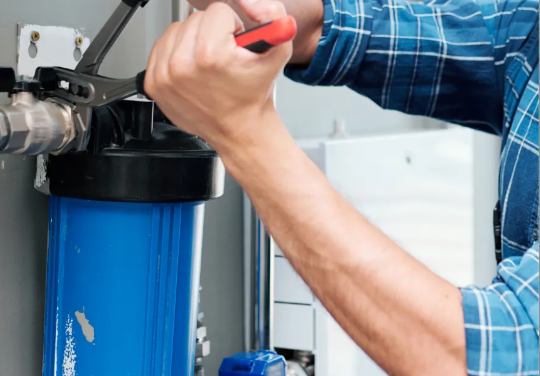 Person in a blue plaid shirt using a wrench to install or fix a blue water filter unit on a wall.