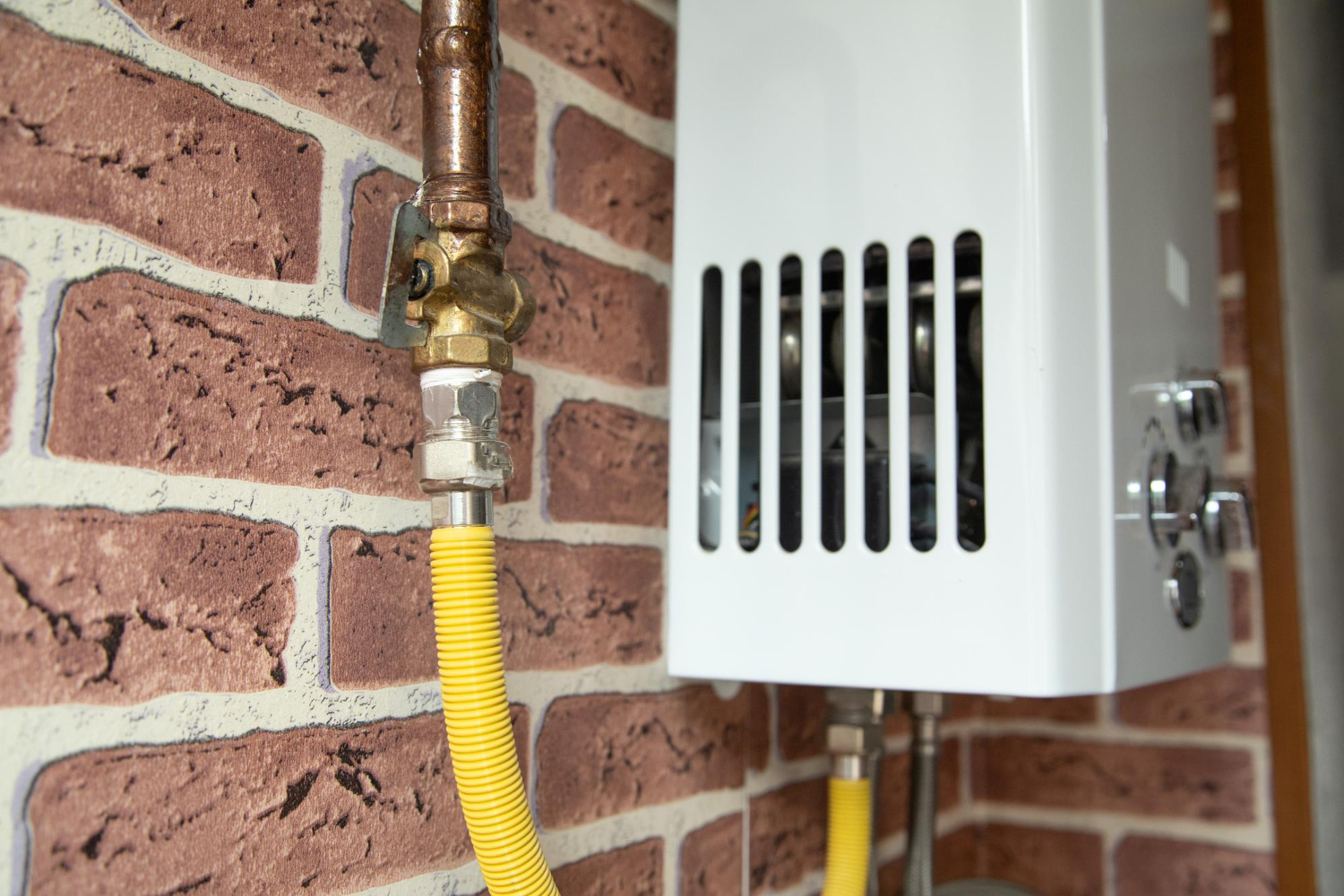 Close-up of a gas water heater with yellow pipe against a brick wall background.