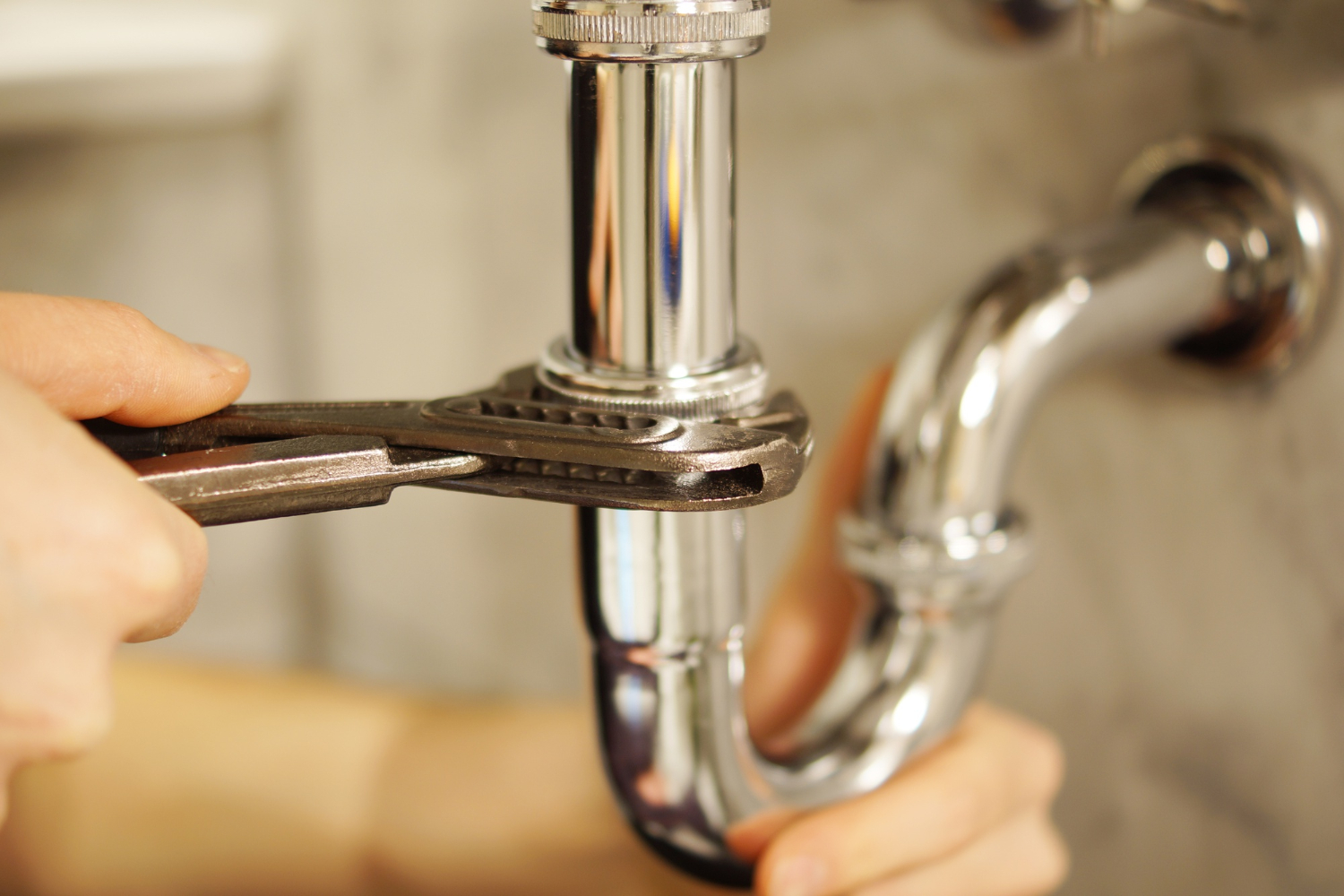 Close-up of hands using a wrench to tighten a shiny metal pipe under a sink.