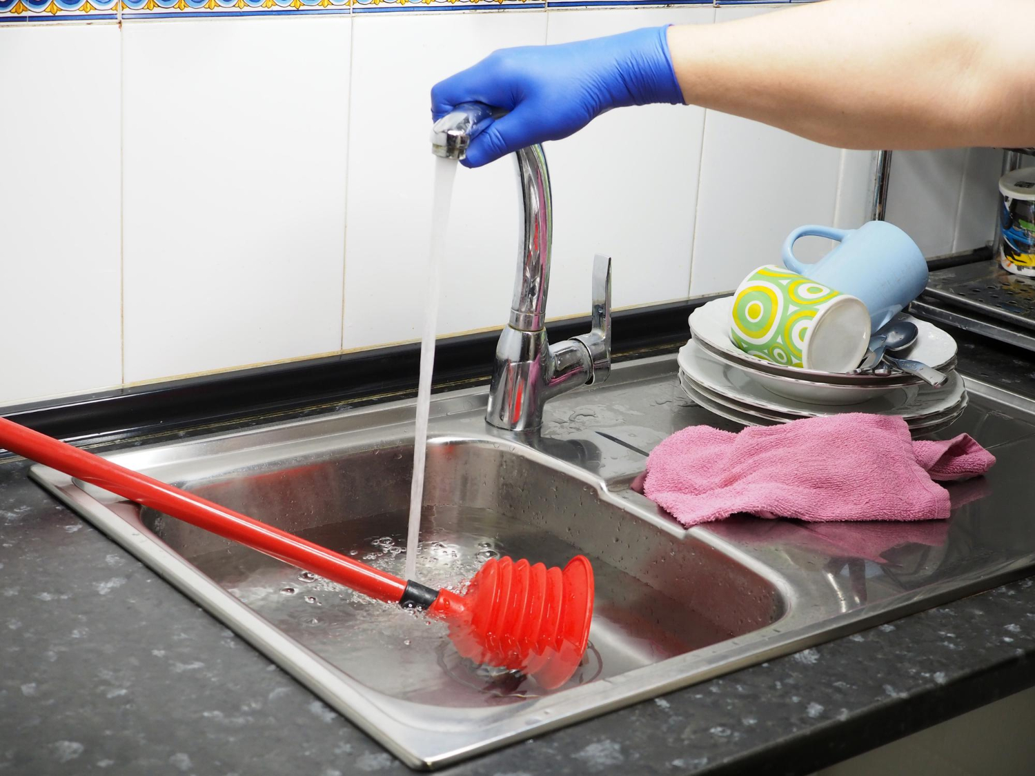 A gloved hand runs water over a plunger in a kitchen sink near stacked dishes and a pink cloth.